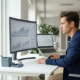 Fachkraft analysiert Finanzdashboard auf großem Monitor am modernen, aufgeräumten Schreibtisch im natürlichen Tageslicht.