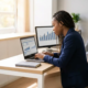 Junge Fachkraft analysiert Finanzdaten auf Laptop und Monitor am modernen Schreibtisch mit natürlichem Licht.
