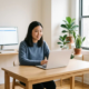 Junge Frau asiatischer Herkunft arbeitet konzentriert an modernem Schreibtisch mit Laptop und Monitor mit Tabellendaten, natürliches Seitenlicht.