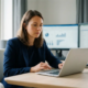 Frau analysiert Finanzdaten auf Laptop an modernem Schreibtisch mit Diagrammen auf Monitoren im Hintergrund, natürliches Seitenlicht.