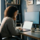Junge Frau arbeitet konzentriert am Laptop in einem aufgeräumten, modernen Büro mit warmem Tageslicht und minimalem Schreibtisch.
