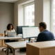 Steuerberater analysiert Finanzdaten auf großem Monitor in modernem Büro, Kollegin arbeitet im Hintergrund am Laptop.